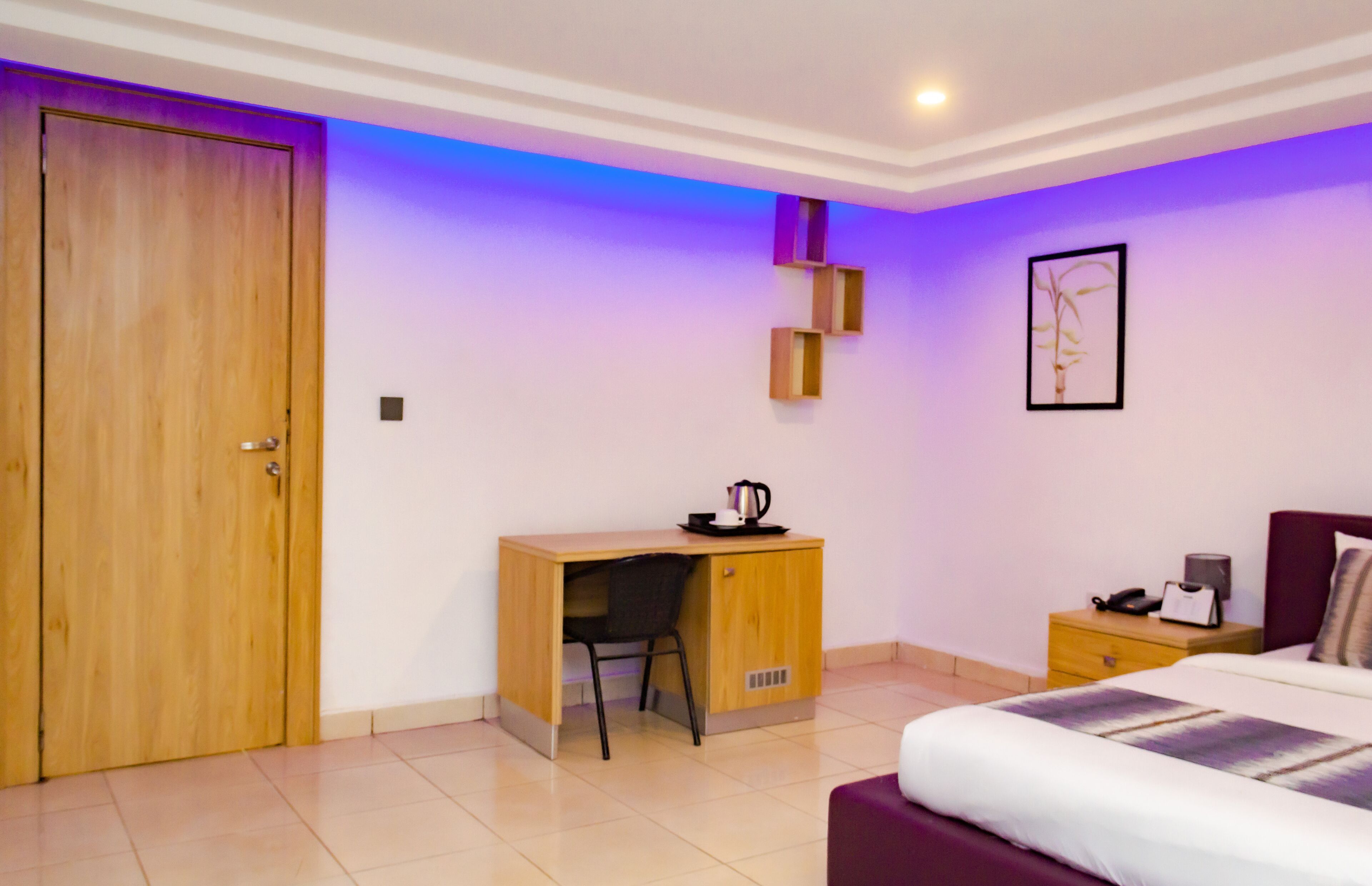 A bedroom with a wooden desk, a chair, a bed with a purple headboard, a nightstand, and a wall-mounted shelf.