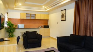 Una sala de estar moderna con un sofá negro, una mesa de centro y un televisor sobre un mueble de madera. Cuenta con una zona de cocina con azulejos anaranjados y armarios de madera.