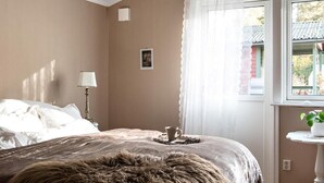 A bedroom with a bed, a bedside lamp, a window with curtains, and a small table with a plant. - Experience the Ultimate in Comfort and Serenity (Stockholms län)