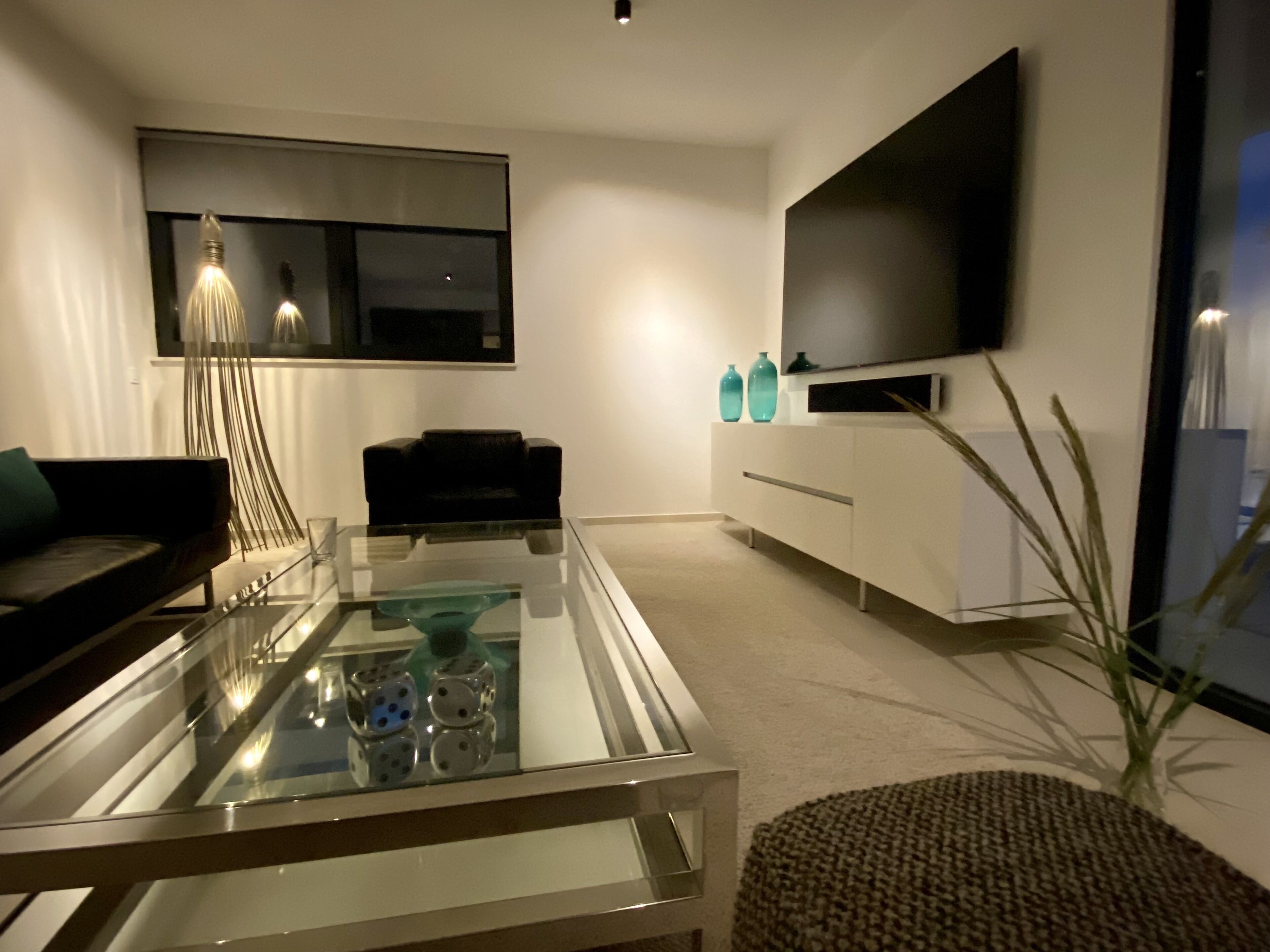 A modern living room with a glass coffee table, a black sofa, a white cabinet, and a large flat-screen TV mounted on the wall.