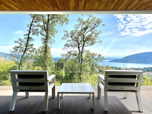 Une terrasse avec deux chaises blanches et une petite table, offrant une vue panoramique sur des arbres et une étendue d’eau.