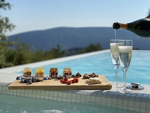Une scène au bord de la piscine, avec une planche en bois sur laquelle sont disposés du fromage et des crackers, deux coupes de champagne et une bouteille dont on verse le contenu.