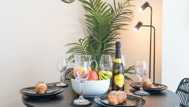 A dining table set with a bowl of fruit, a bottle of champagne, and pastries. - Afton (Lancaster)