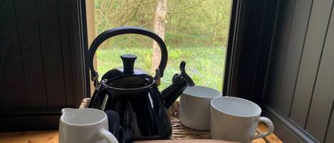 A black teapot with a lid, two white mugs, and a book titled "The Fall of the Sparrow" on a woven mat by a window with a view of trees.