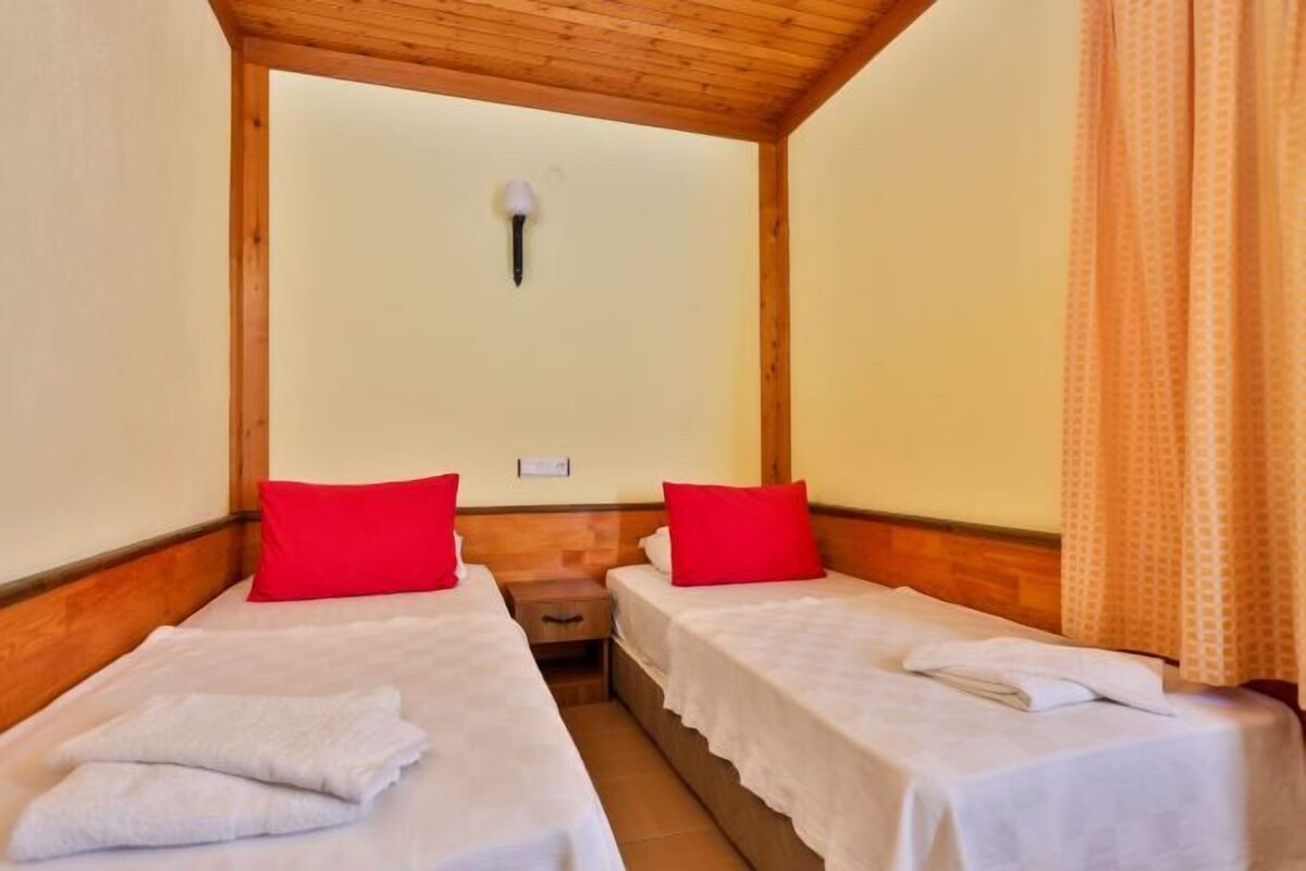 A room with two single beds, each with a red pillow and a white towel. There is a wooden headboard, a small bedside table, and a wall-mounted light fixture.