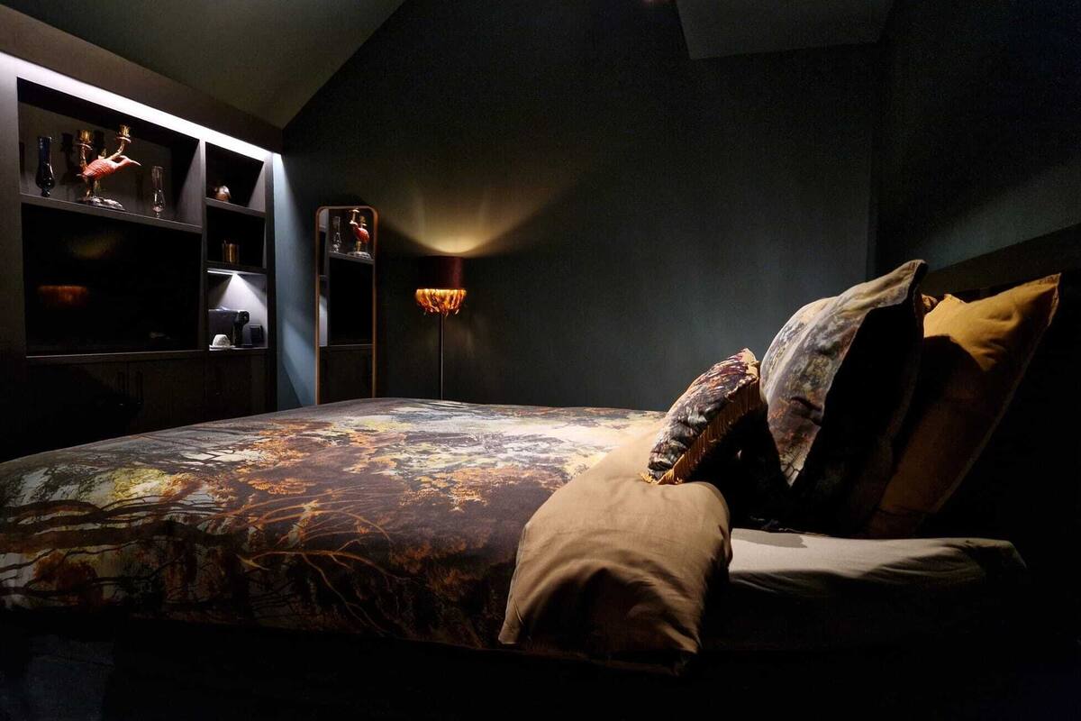 A bedroom with a bed, pillows, a bedside table, a lamp, and a shelf with decorative items.