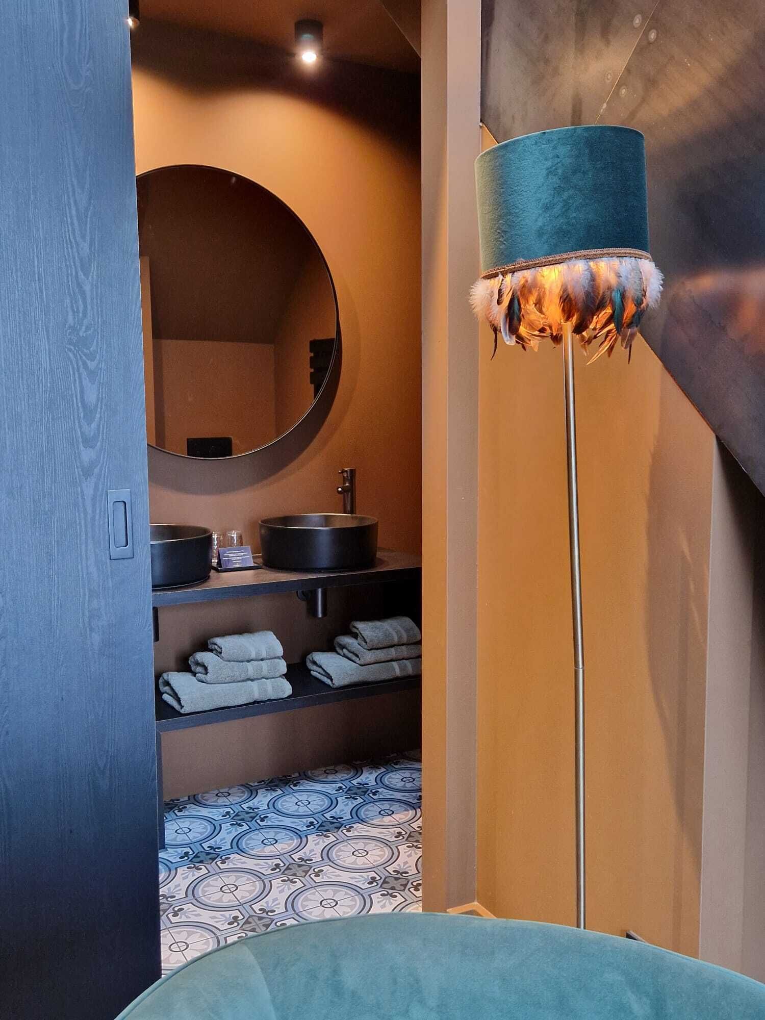 A modern bathroom with a round mirror, a sink with a black basin, and a teal floor lamp with a feathered shade.