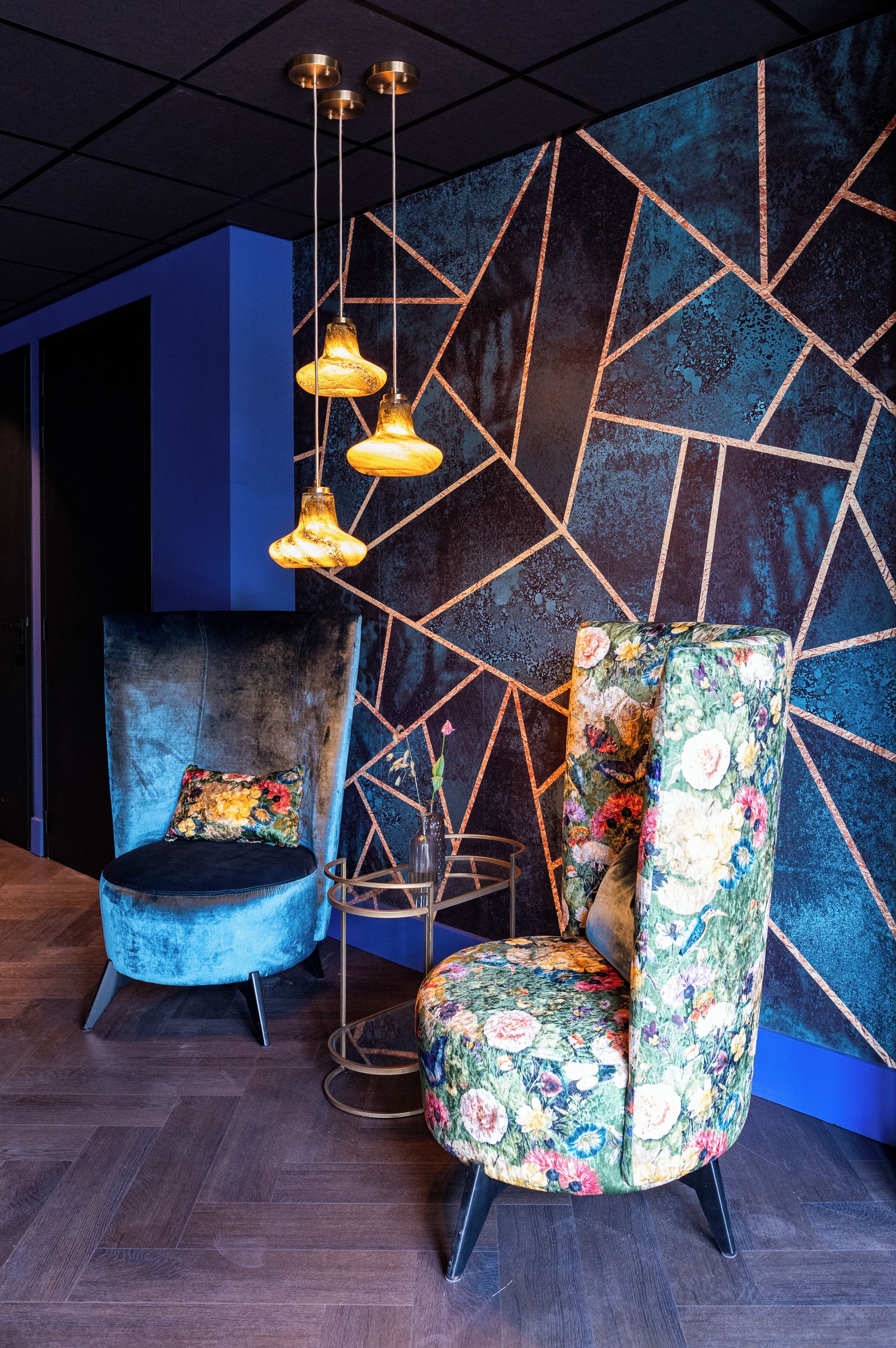 Una habitación moderna con un diseño geométrico en la pared, un sillón de terciopelo azul y un sillón floral, además de lámparas colgantes.