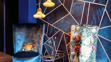 Una habitación moderna con un diseño geométrico en la pared, un sillón de terciopelo azul y un sillón floral, además de lámparas colgantes.