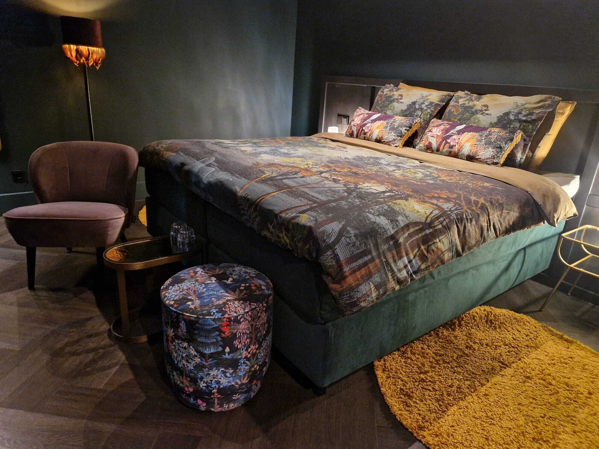 Un dormitorio moderno con una cama grande, una silla, un otoman redondo y una lámpara de pie.