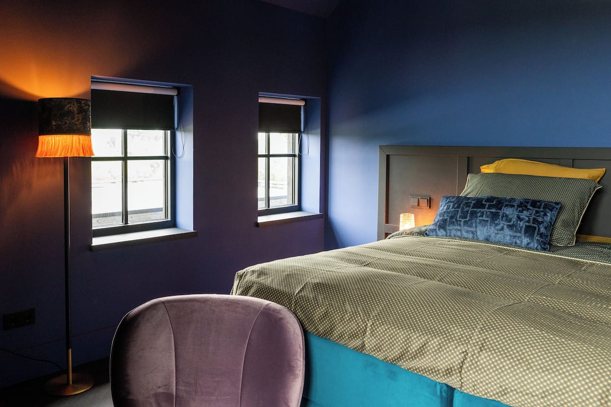 A bedroom with a bed, two windows, a floor lamp, and a chair.