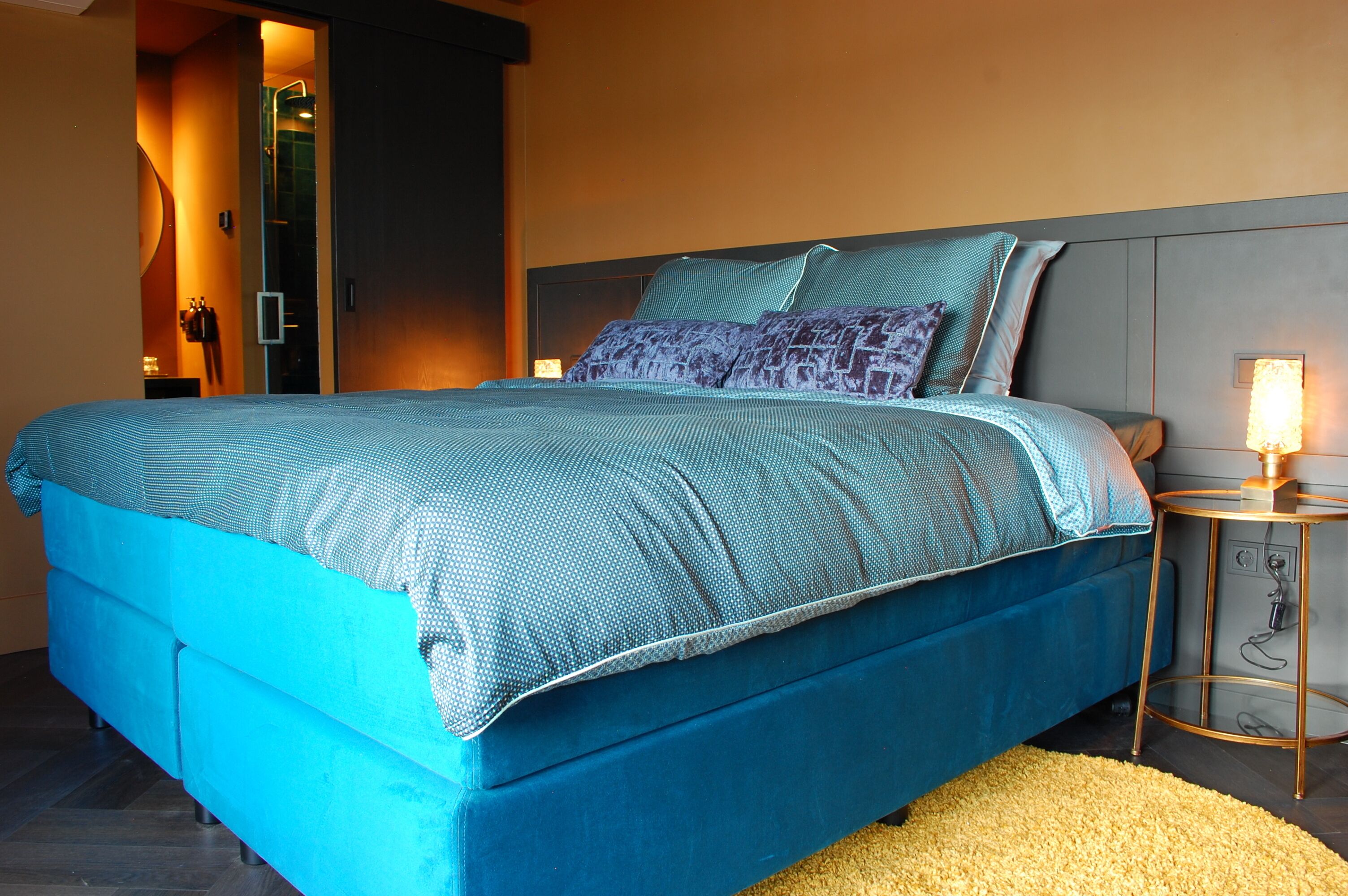 Una habitación de hotel moderna con una cama grande, una mesita de noche pequeña y una zona de ducha con mampara de cristal.