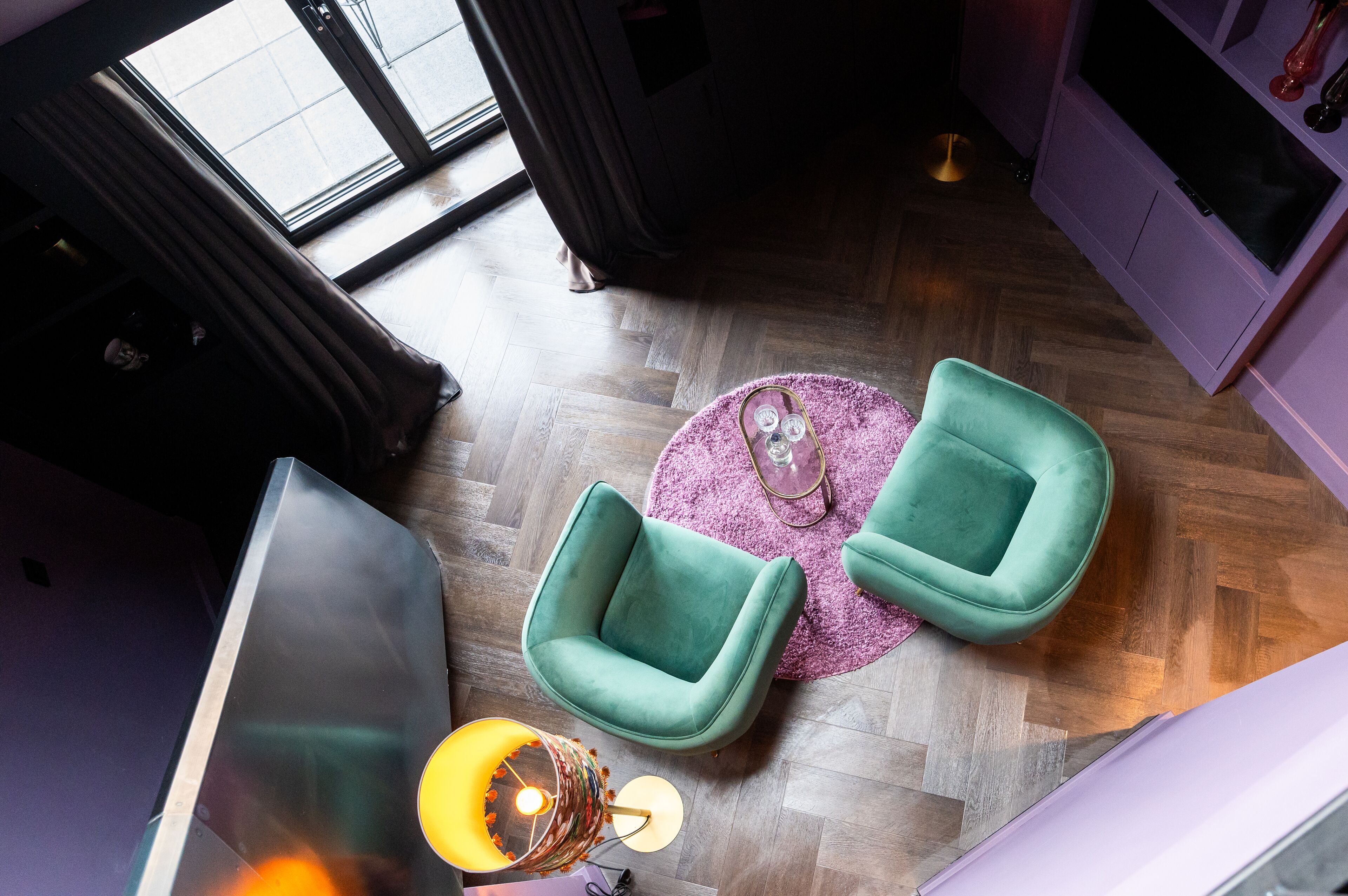 Ein modernes Wohnzimmer mit Holzboden, zwei grüne Sessel, einem runden Teppich in Rosa und einem Glastisch.