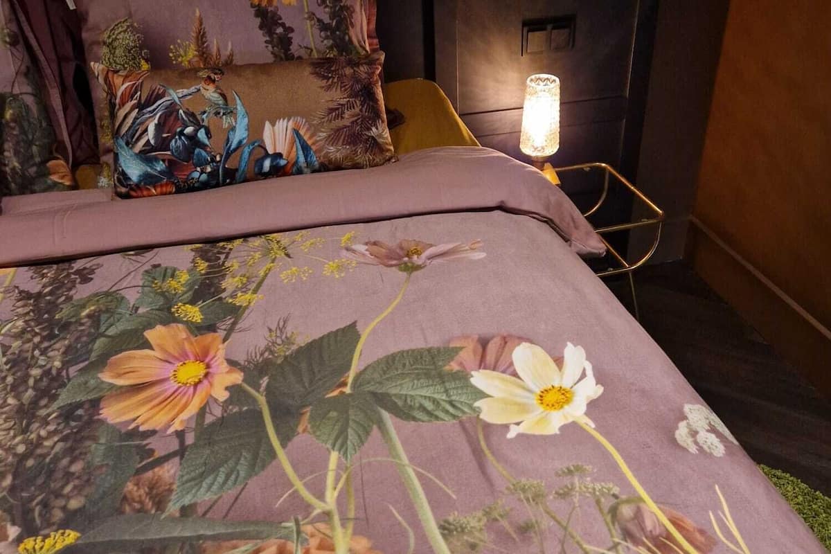 A bed with floral bedding, a nightstand with a lamp, and a chair in a dimly lit room.