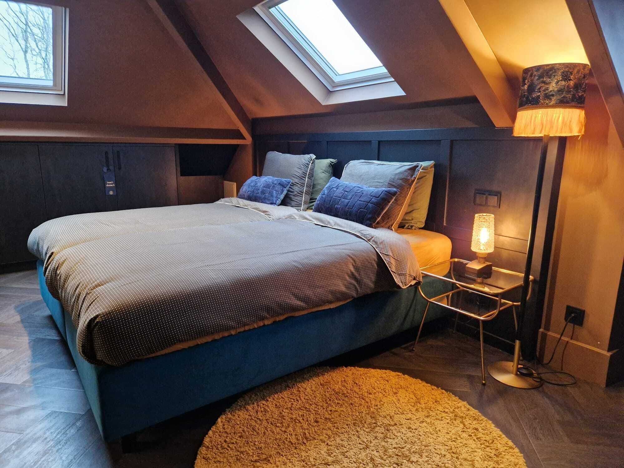 A bedroom with a sloped ceiling, a bed with a patterned bedspread, multiple pillows, a bedside lamp, and a round rug.