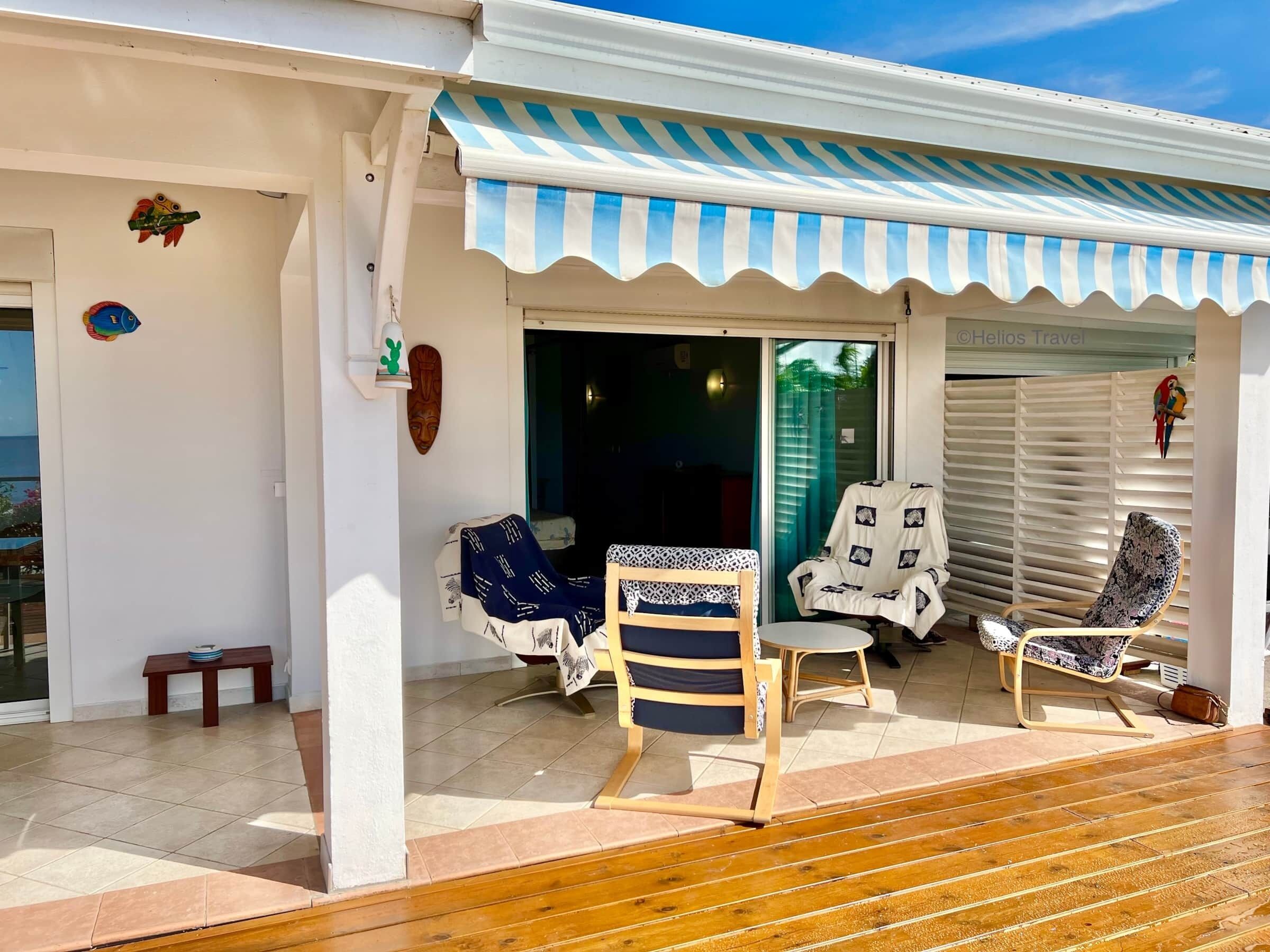 Un espace extérieur abrité, aménagé avec deux chaises et un banc, une petite table, et une porte donnant accès à un espace intérieur.