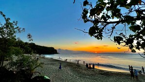 Ein Strand mit Menschen, die laufen und stehen, Bäumen und einem Sonnenuntergang über dem Wasser.