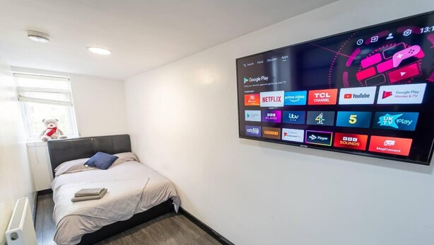 A modern hotel room with a large digital display on the wall, a neatly made bed, a small rug, and a window with a teddy bear. - Studio Close to Selly Oak Train Station (Birmingham)