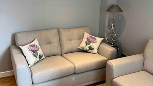 A living room with a beige sofa, two decorative pillows, a framed botanical print on the wall, and a floor lamp. - Inverness Highland Apartment (Inverness)