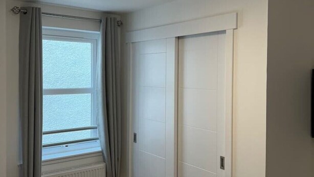 A bedroom with a bed, a window with curtains, a radiator, a ceiling light fixture, and a door. - Inverness Highland Apartment (Inverness)