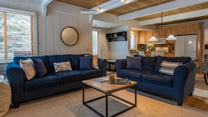 Ein modernes Wohnzimmer mit zwei dunkelblauen Sofas, einem Holztisch und einer Küche im Hintergrund.