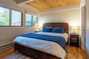Une chambre à coucher avec une tête de lit en bois, un grand lit avec une literie bleue et blanche, deux lampes de chevet et une vue sur l’extérieur par deux fenêtres.