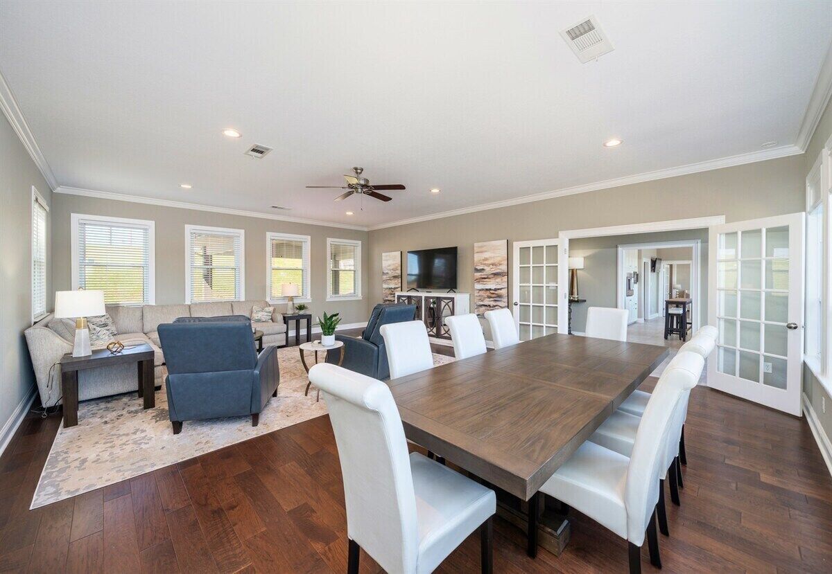 A spacious living room with a large wooden dining table, white chairs, a sofa, and a ceiling fan.