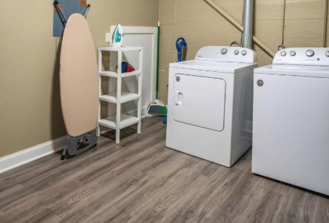 A laundry room with a white shelf, two white washing machines, and a beige ironing board.