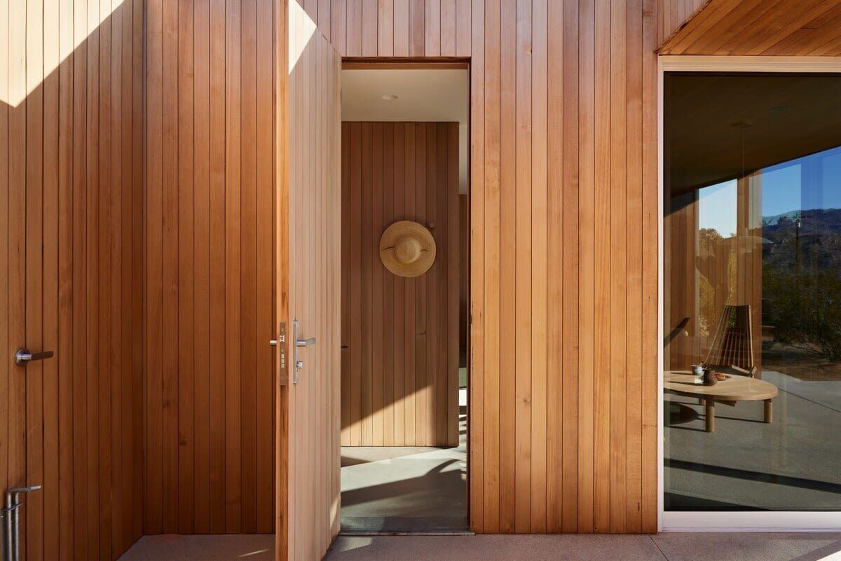 Pared exterior de madera con una puerta que da a una habitación, donde se ve un sombrero en la pared y una mesa con sillas afuera.