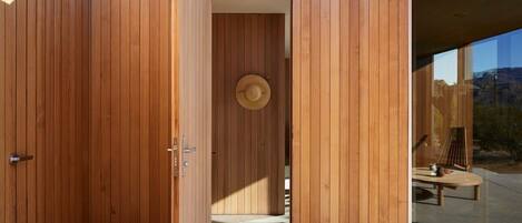 Pared exterior de madera con una puerta que da a una habitación, donde se ve un sombrero en la pared y una mesa con sillas afuera.