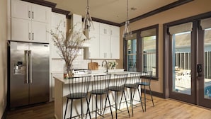 A modern kitchen with a stainless steel refrigerator, white cabinets, a central island with bar stools, and a large window with a view of the outdoors. - Chic, Marigny Retreat | Heated Pool (New Orleans)