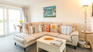 A living room with a beige sofa, a coffee table with books and lemons, a painting on the wall, and a floor lamp. - Gull Cottage (Lowestoft)
