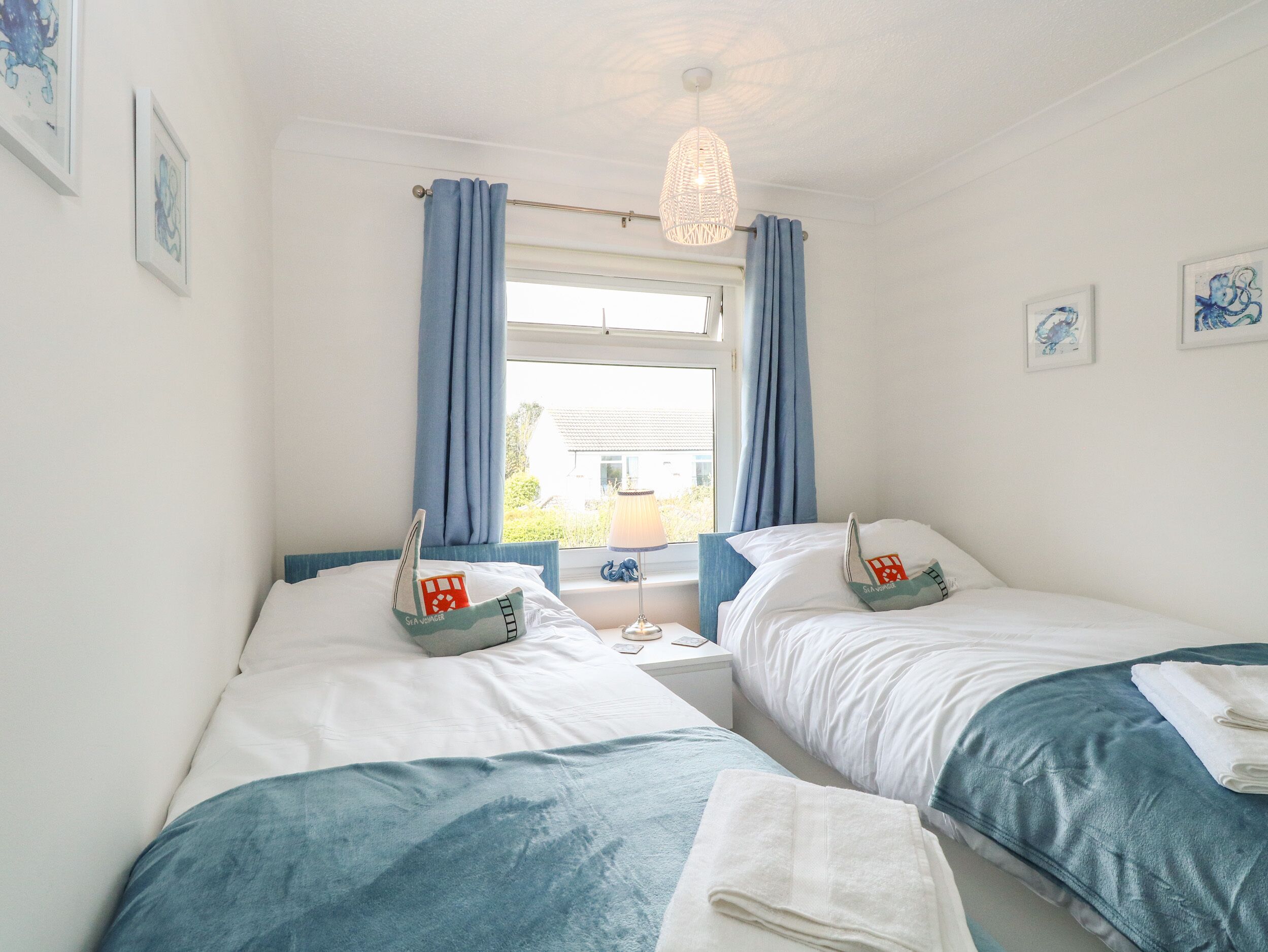 A bedroom with two beds, a window with curtains, a bedside table with a lamp, and artwork on the walls.