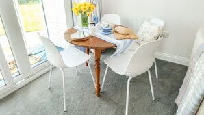 A dining area with a round wooden table, white chairs, a vase of yellow flowers, and a view of the outdoors through large windows. - Gull Cottage (Lowestoft)
