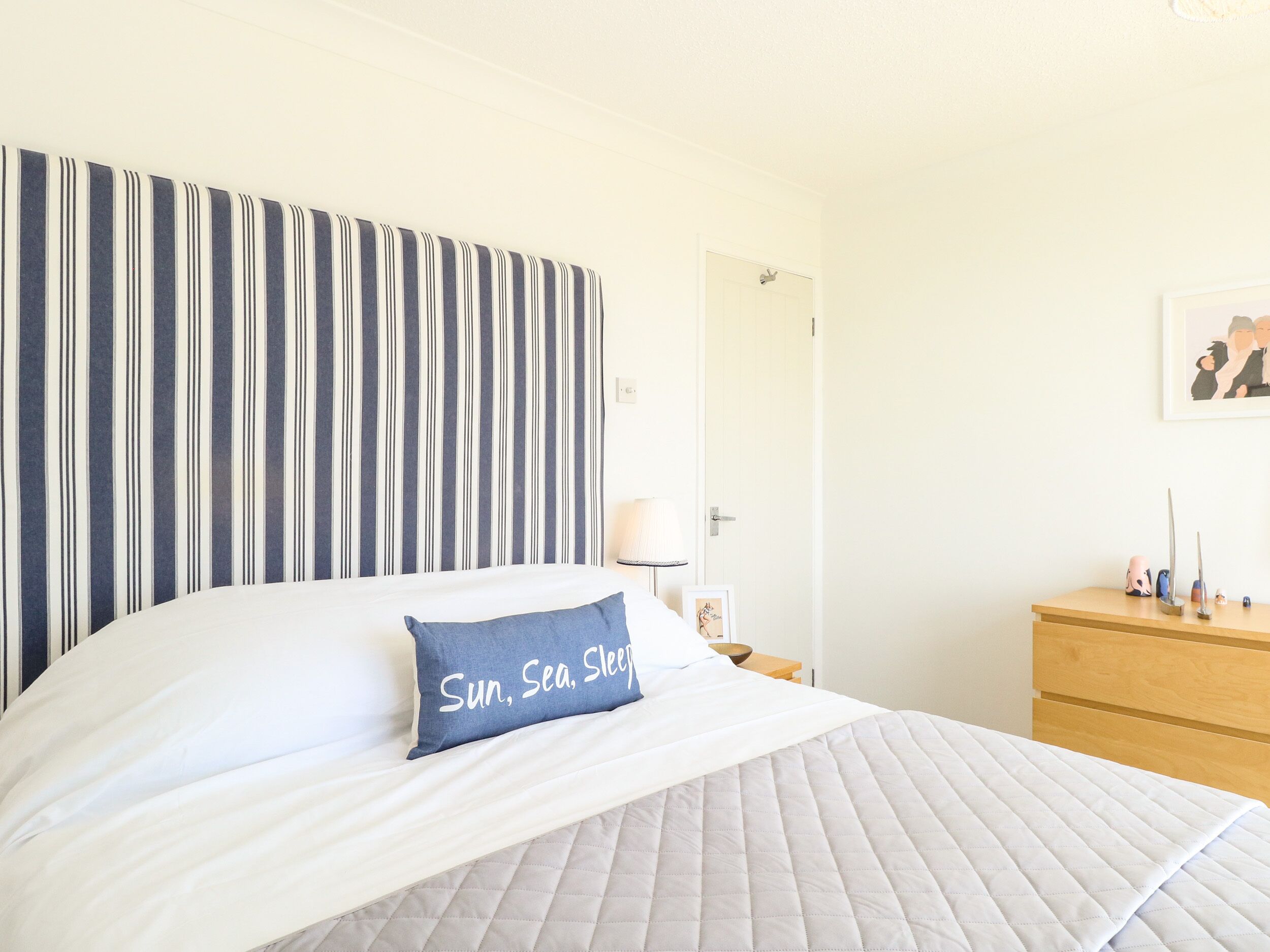 Ein ordentlich bezogenes Bett mit einem Kissen, auf dem "Sun, Sea, Sleep" steht, in einem Zimmer mit gestreiften Vorhängen und einer Holzkommode.