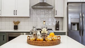 Ett kök med vita skåp, en köksfläkt i rostfritt stål och ett kylskåp. På bänkskivan står ett bricka med glas och flaskor.