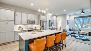 A modern kitchen with white cabinets, a central island with bar stools, a dining area with a sofa and table, and a large window with a view. - 4BR North Nashville Retreat | Rooftop Deck & Grill (Nashville)