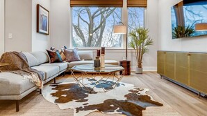 A modern living room with a sectional sofa, a glass coffee table, a cowhide rug, a potted plant, and a large window with a view of trees. - 4BR North Nashville Retreat | Rooftop Deck & Grill (Nashville)