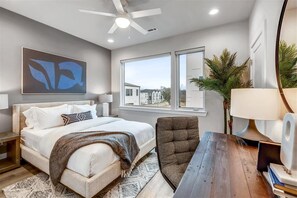 Une chambre moderne comprenant un lit, un bureau, une chaise, un ventilateur de plafond, une fenêtre avec vue et un grand tableau abstrait.