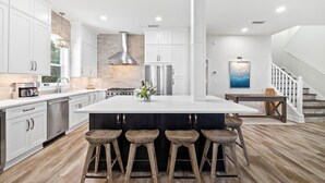 A modern kitchen with white cabinetry, a central island with stools, a stainless steel refrigerator, and a brick accent wall. - Gulf Views | Pool | Close to Beach | Elevator (Santa Rosa Beach)