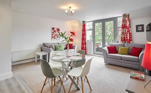 A modern living room with a grey sofa, red and yellow cushions, a glass dining table with chairs, and a red lamp.