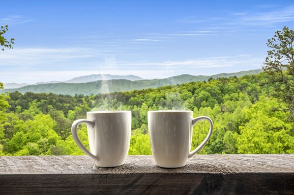 Deux tasses blanches sont posées sur une étagère en bois, avec en arrière-plan une chaîne de montagnes boisée.