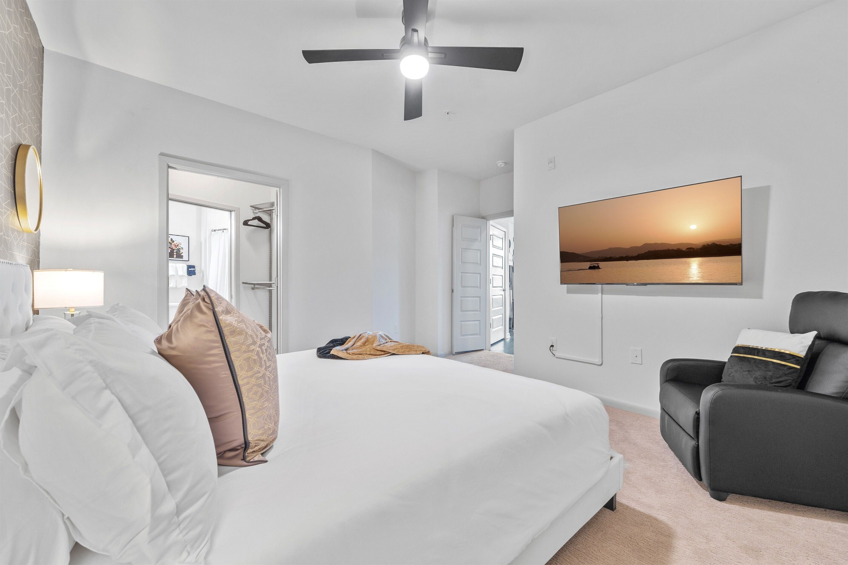 A hotel room with a bed, a TV mounted on the wall, a ceiling fan, and a view of a sunset over a body of water.