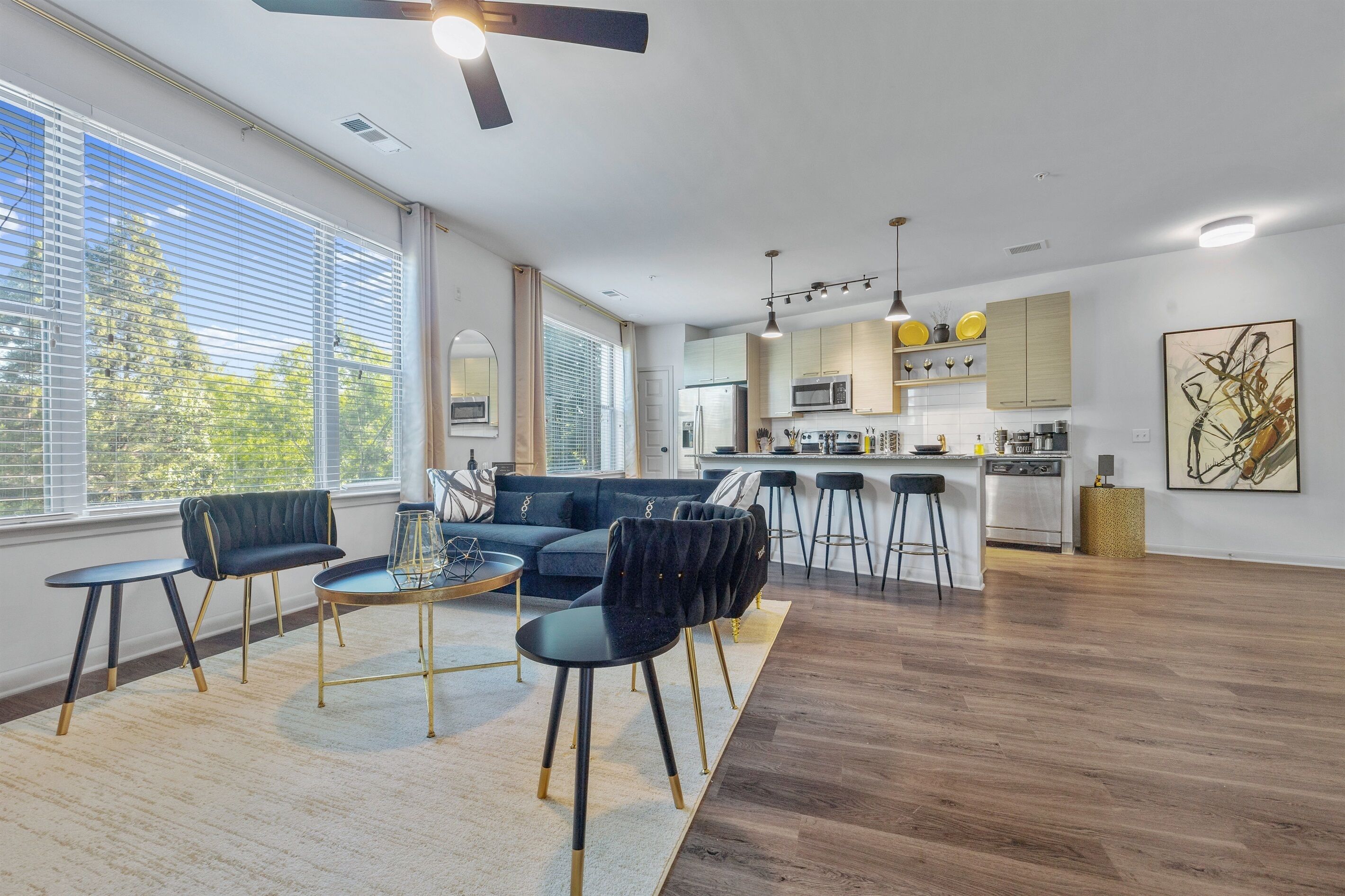 A modern living room with a sofa, chairs, and a coffee table. There is a kitchen area with a microwave, stove, and bar stools.