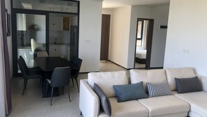 A modern living room with a sofa, a dining area with a table and chairs, and a kitchen visible through a glass door. - Phoenix Villa (Kigali)