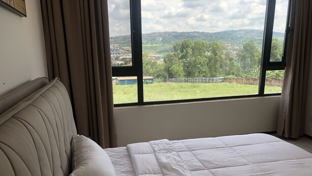 A hotel room with a bed, a window with a view of a green landscape, and curtains. - Phoenix Villa (Kigali)