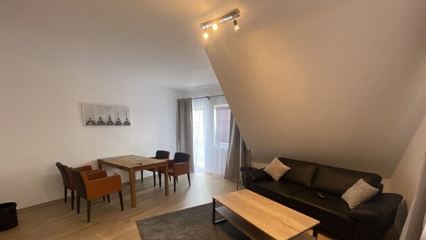 Una sala de estar moderna con una mesa de comedor de madera, cuatro sillas, un sofá negro y una alfombra gris.