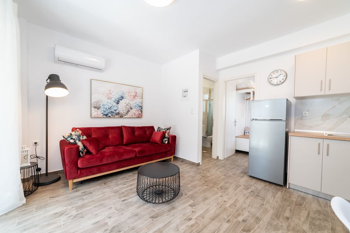 A compact living space with a red sofa, a round black table, a kitchen area with a fridge, and a clock on the wall.