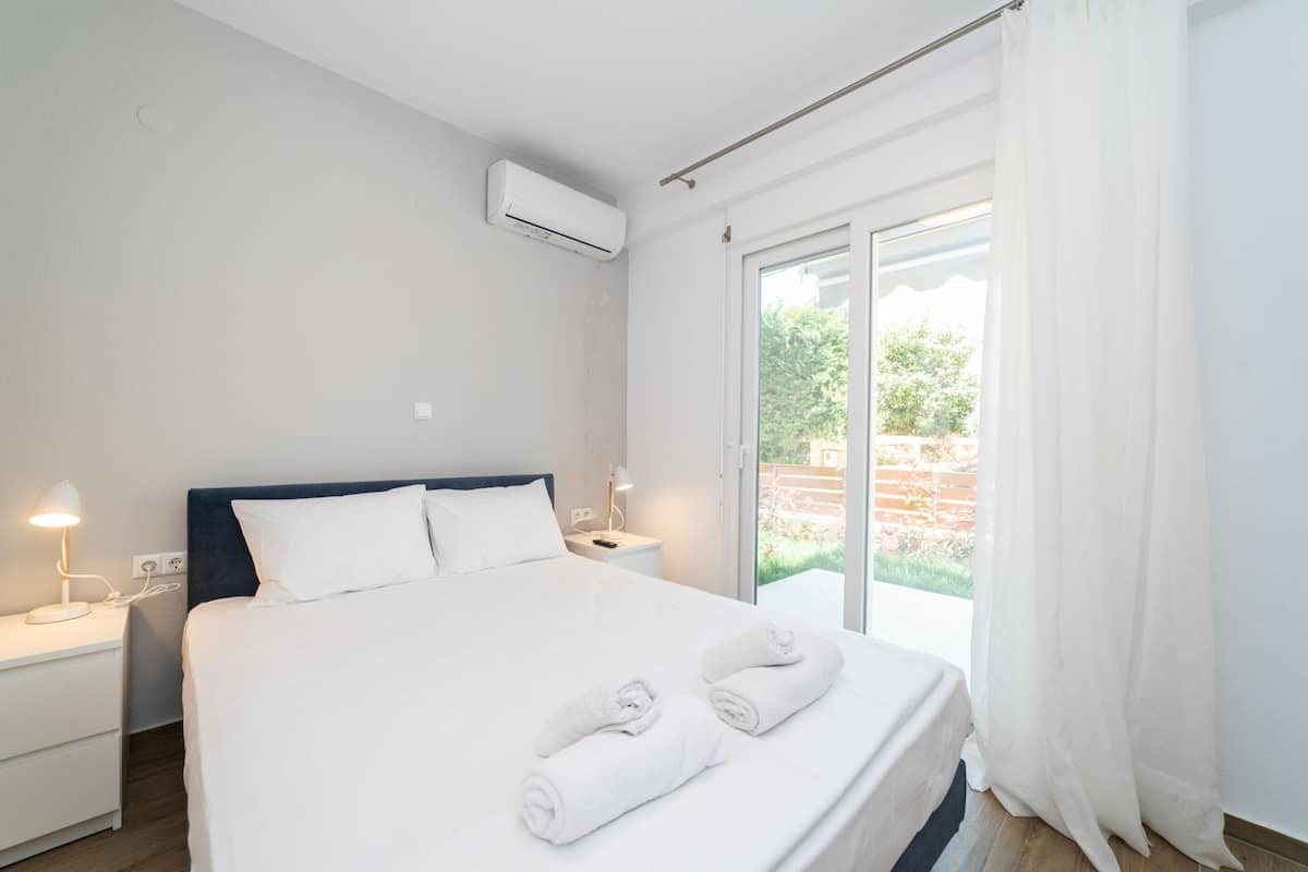 A modern hotel room with a large bed, two bedside tables, an air conditioning unit, and a sliding glass door leading to an outdoor area.