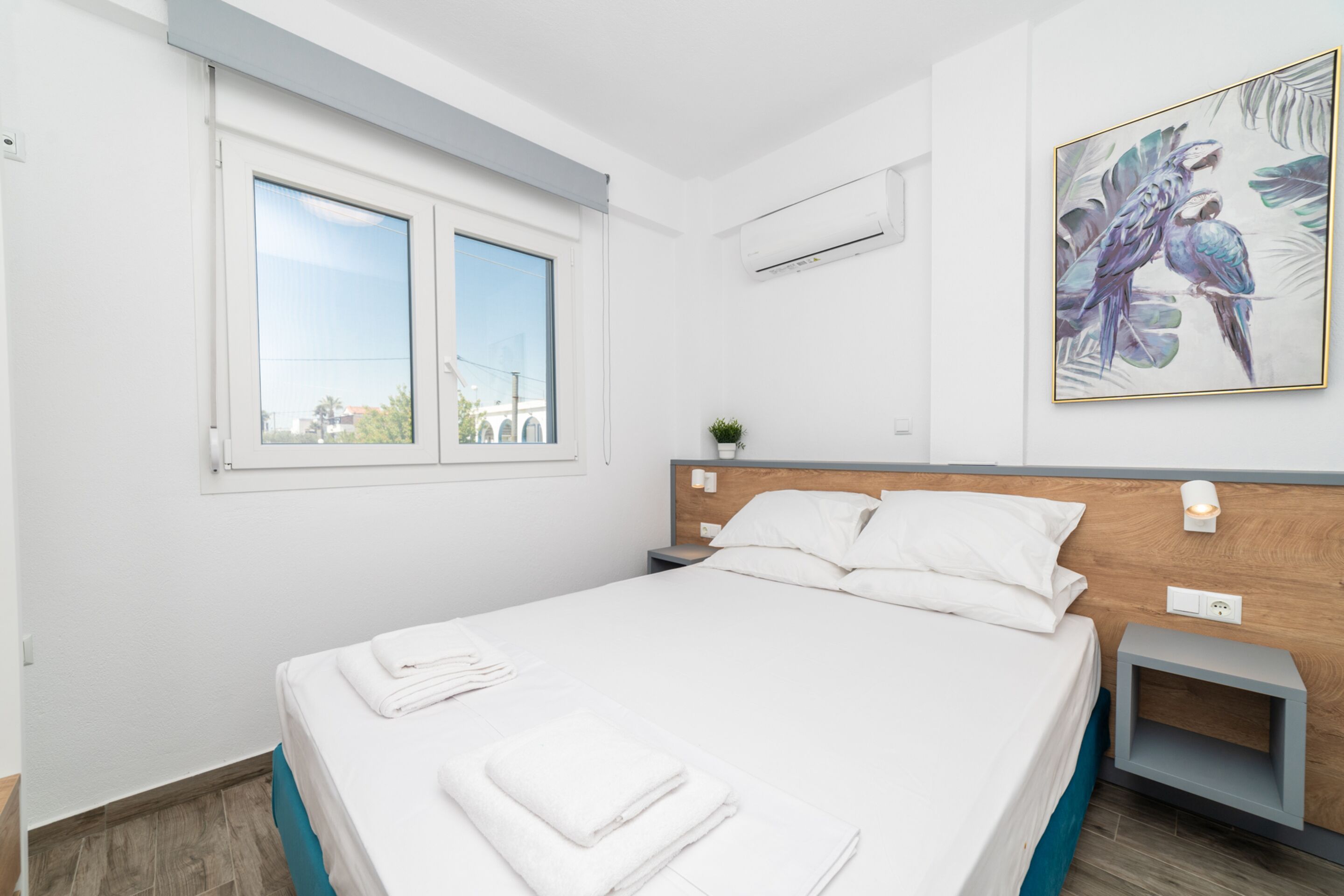 A modern hotel room with a large bed, a small bedside table, a nightstand, a window, and an air conditioning unit.